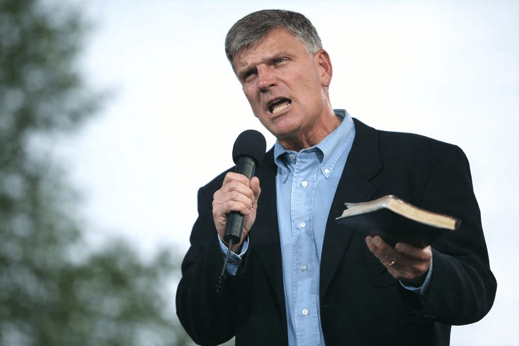 Close up of Franklin Graham outdoors holding a microphone in one hand and a Bible in the other while addressing a crowd.