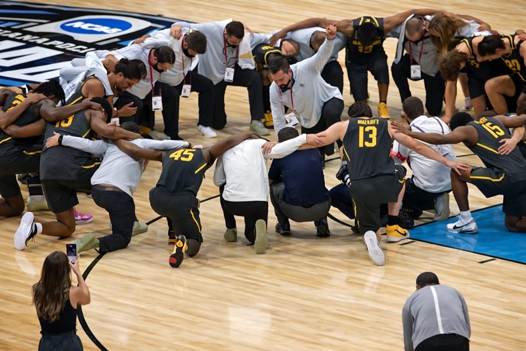 Baylor basketball players and coaches kneel in a linked circle at center court in prayer after a game on an NCAA tournament hardwood court.