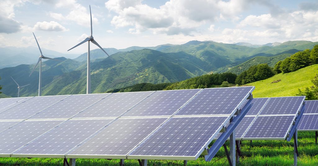 Solar panels and wind turbines on a green mountainside under a bright, cloudy sky.
