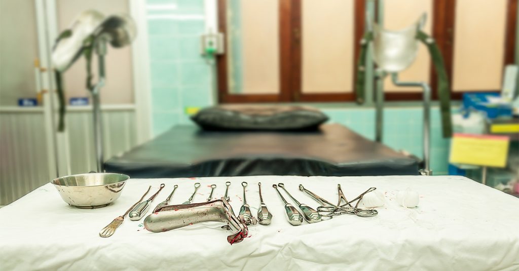 Surgical instruments with visible blood stains arranged on a draped medical table in an exam room, with a gynecological procedure chair in the background.
