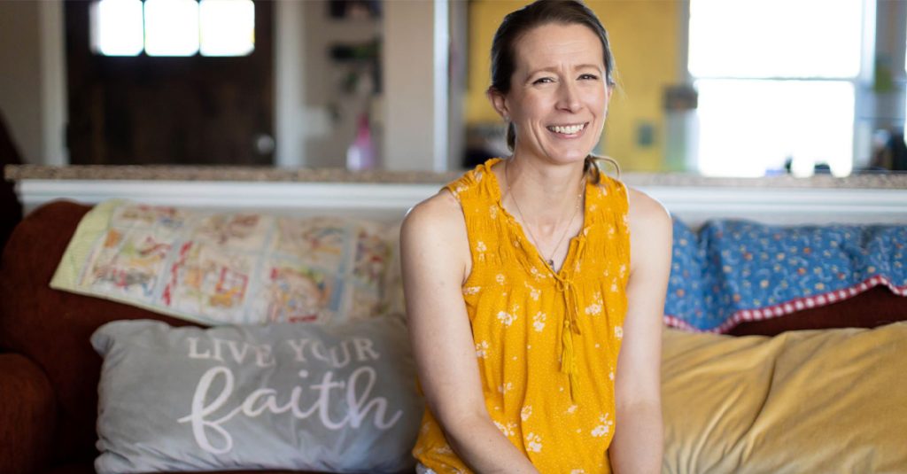 Jessica Bates sits in her home in front of pillows that reads, "Live Your Faith."