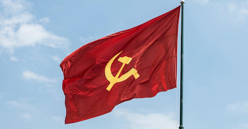 China communist flag with a yellow hammer and sickle flying on a flagpole against a blue sky.