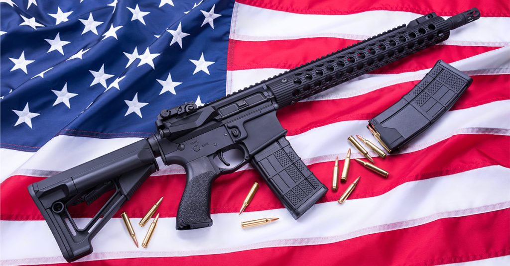 Black AR-15-style rifle, two detachable magazines, and loose ammunition arranged on an American flag.