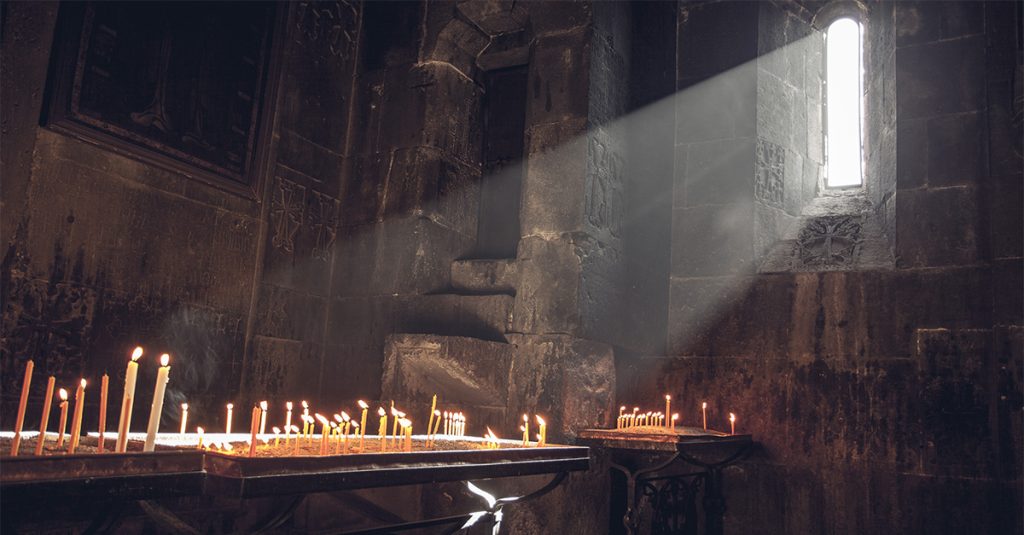Sunlight streams through a narrow window inside a stone church as candles burn on a table below.
