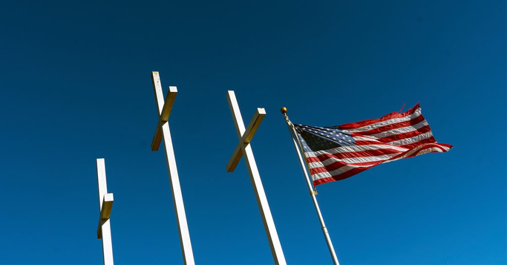 Three white crosses beside a waving American flag against a clear blue sky.