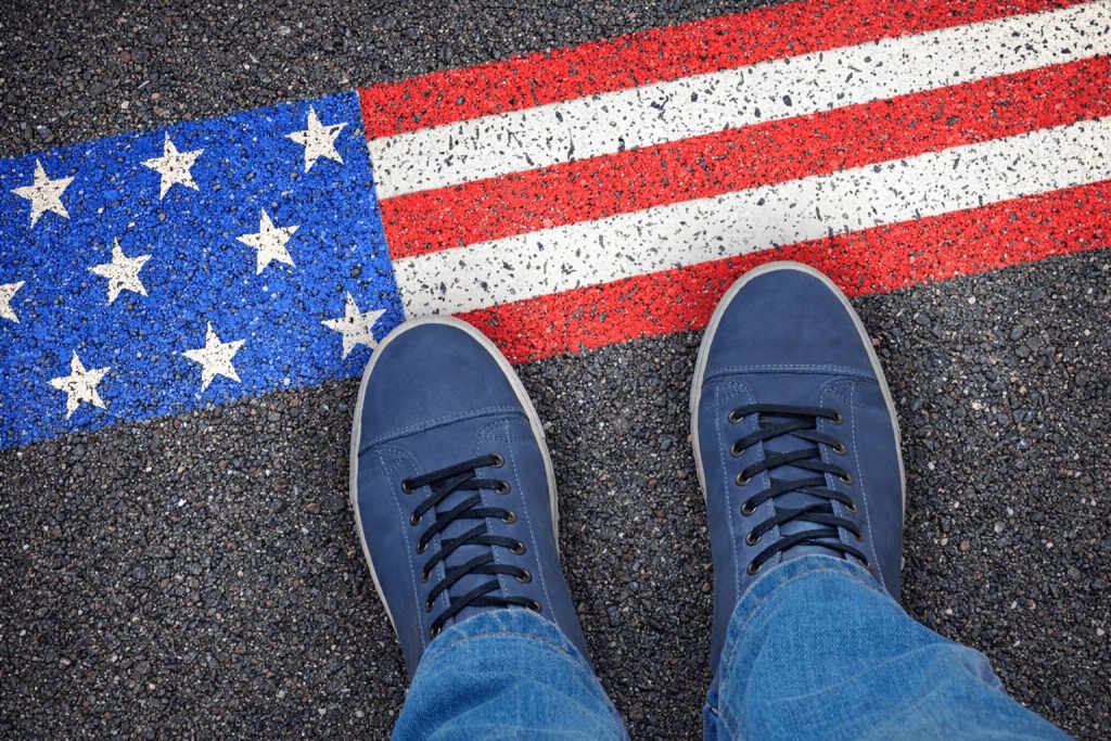 Person standing in blue shoes on an American flag painted across asphalt, symbolizing the line between legal immigration, national identity, and the rule of law.