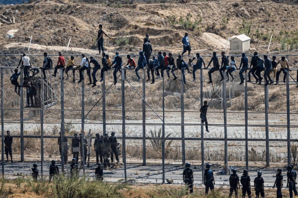 Dozens of migrants climb and sit atop a tall border fence lined with razor wire while officers stand below in a dry, rugged border landscape.