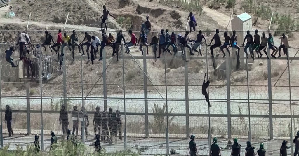 A large group of people sit and climb on a tall border fence while officers stand below on the other side.
