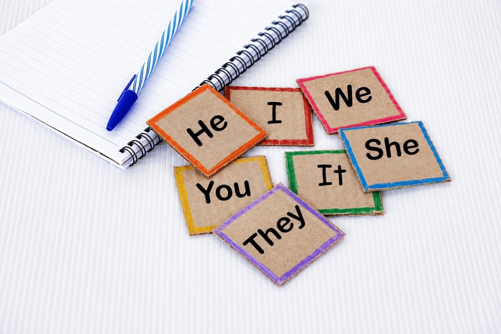 Pronoun cards labeled he, she, they, we, I, you, and it arranged beside a notebook and pen, representing debate over compelled pronoun use in schools.