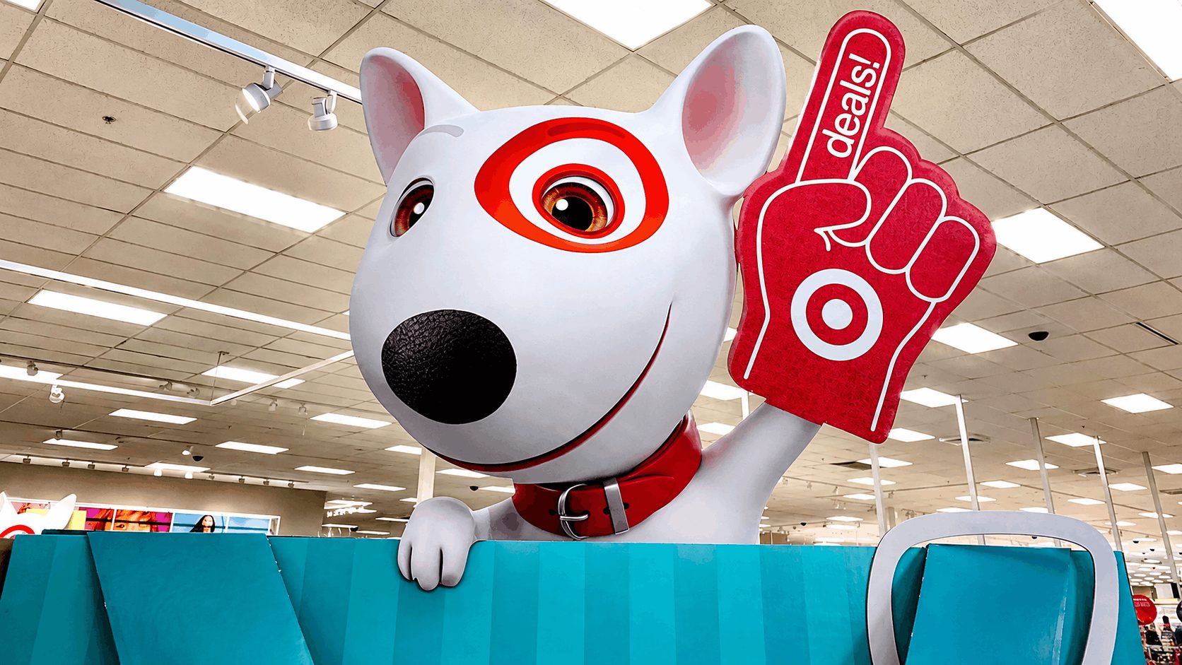 Target’s Bullseye mascot holds a foam deals sign above a retail display inside a brightly lit store.