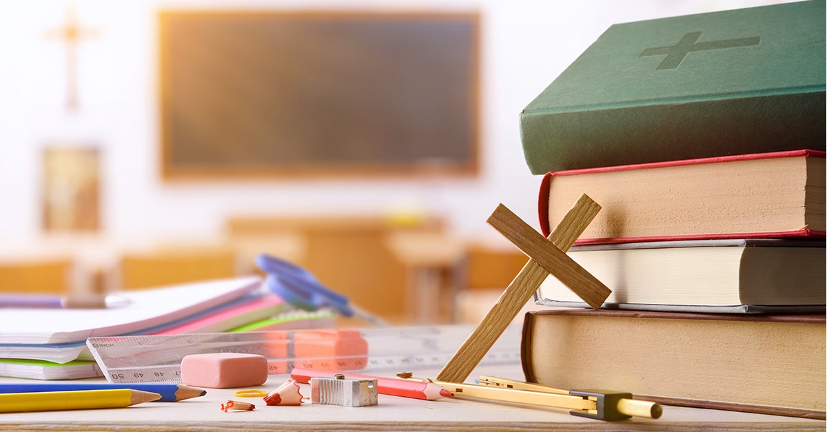 Small wood cross and Bible next to school supplies inside a school classroom