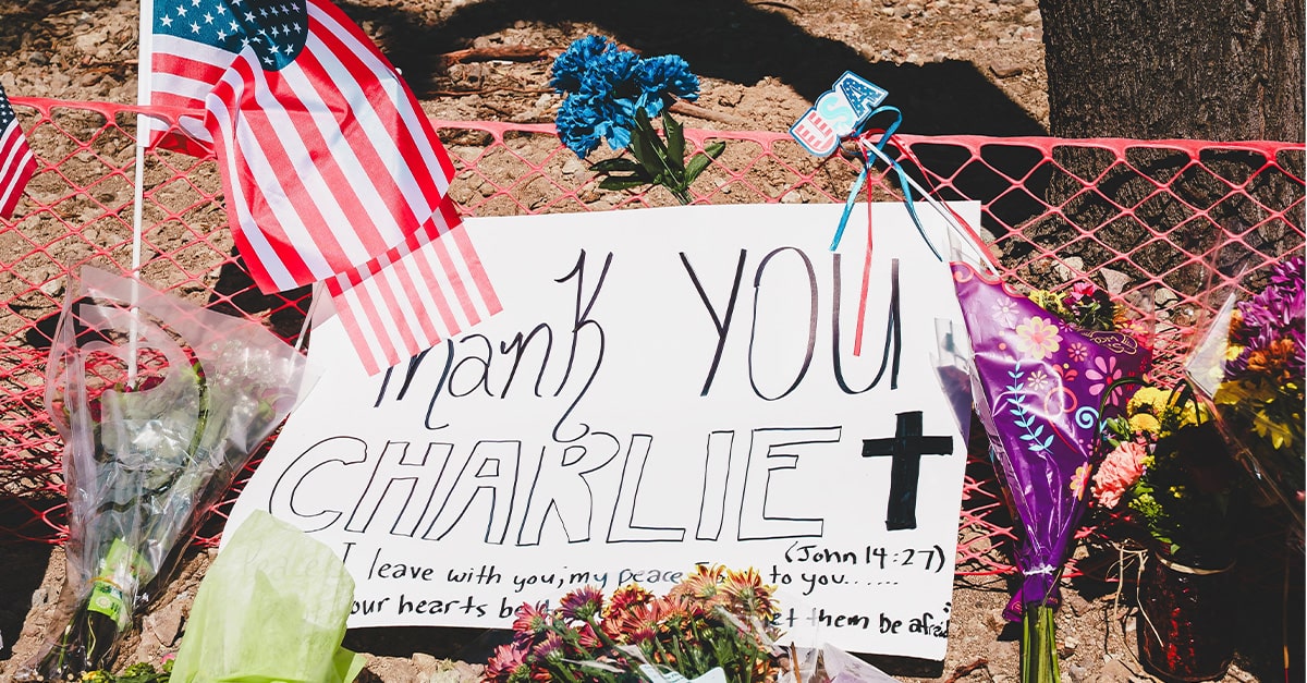 A memorial to Charlie Kirk includes an American flag, flowers, and a poster that reads, "Thank you Charlie," followed by a drawing of a cross and the words to the Bible verse John 14:27.