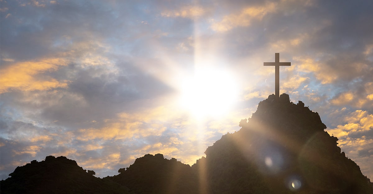 As cross stands in the foreground, the sun rises and shines light in a dark sky.