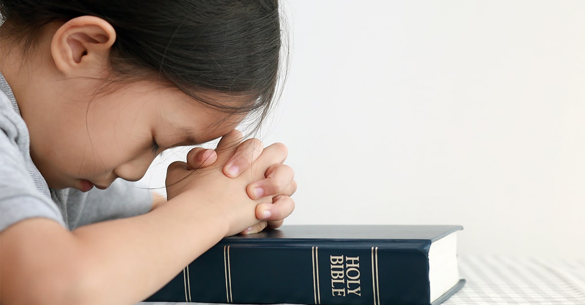 A young girl prays with hands folded on a Bible.