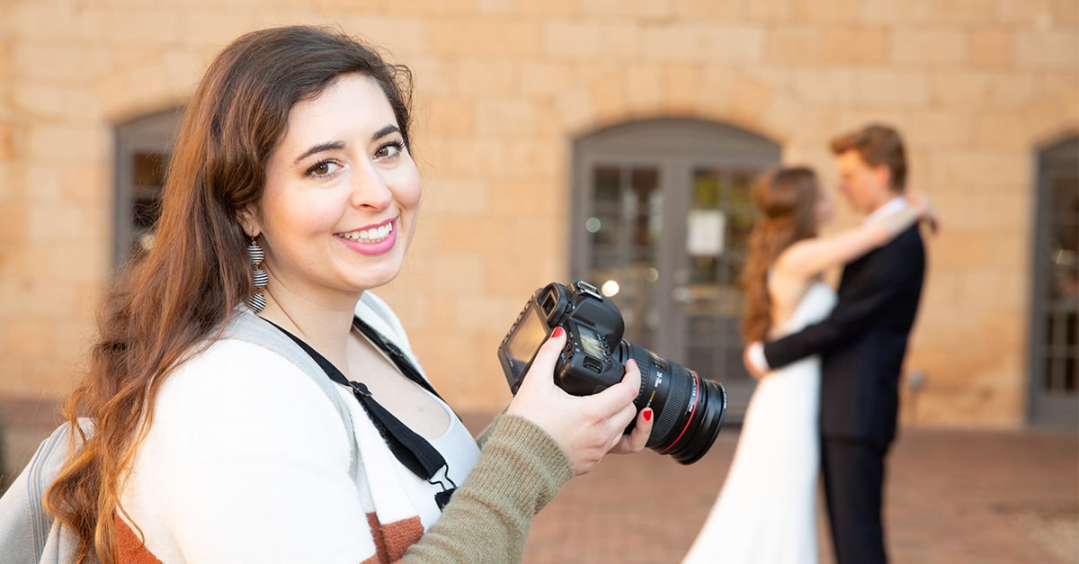 Christian wedding photographer Chelsey Nelson holds her camera and smiles while behind her a man and woman dressed for their big day pose for pictures.