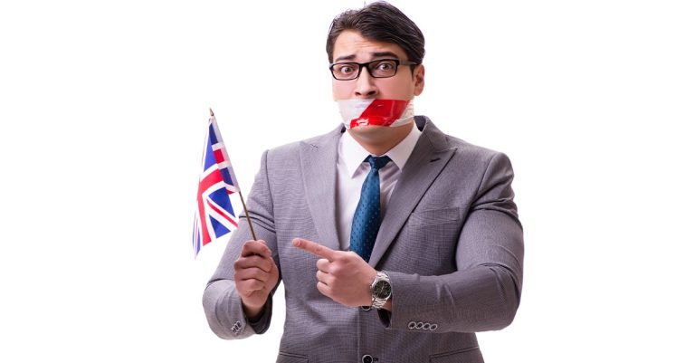 A British Man with mouth taped holds a small Union Jack flag and points to it.