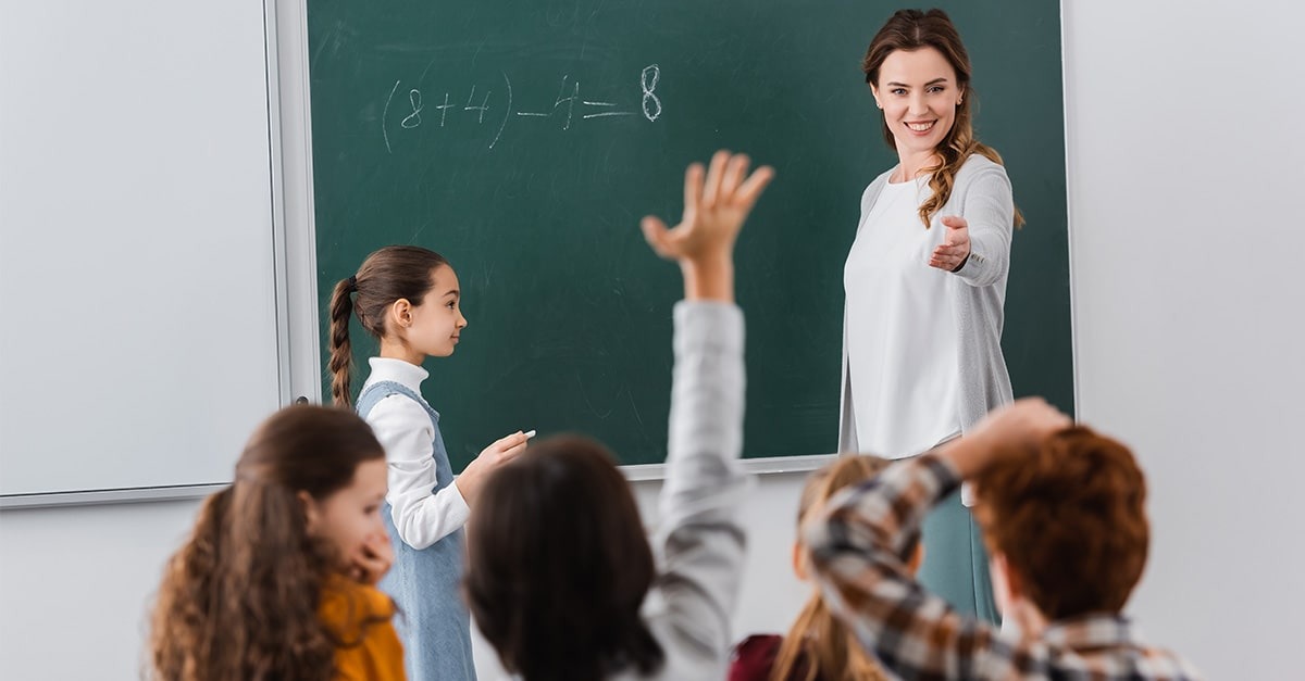 A young teacher stands in front of a blackboard with a math problem and calls on a student raising his hand to answer.