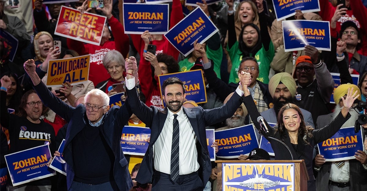 Bernie Sanders and AOC hold New York City Mayor-elect Zohran Mamdani's hands up in a victory pose while on the campaign trail in front of a crowd of supporters holding signs calling for free rent and universal childcare.