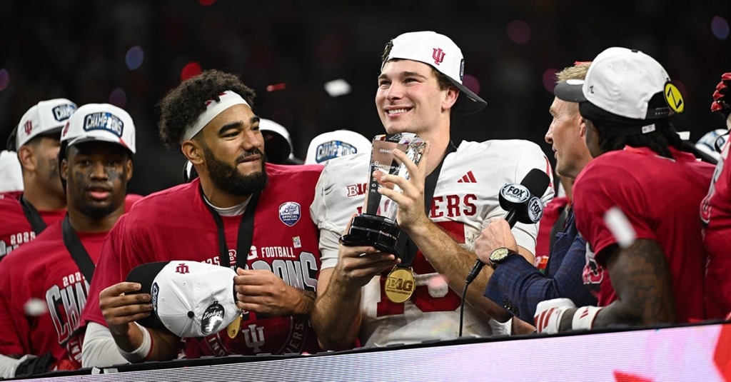 Indiana Quarterback Fernando Mendoza is seen holding the MVP trophy and smiling at the sky while his teammates celebrate around him.
