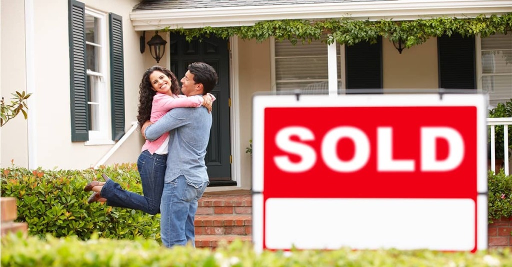 Couple hugging outside a house with a large “SOLD” sign in front.