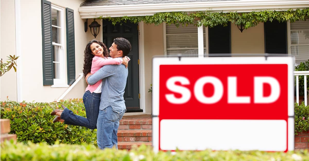 Couple hugging outside a house with a large “SOLD” sign in front.