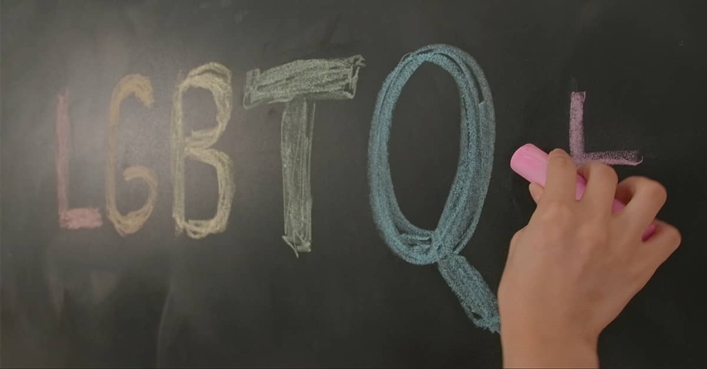 A hand writes “LGBTQ+” in colored chalk on a classroom chalkboard.