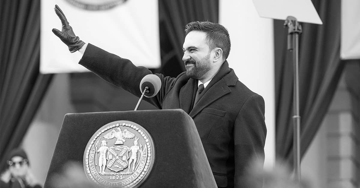 Black-and-white photo of Zohran Mamdani at a podium with the New York City seal, raising his hand in greeting.