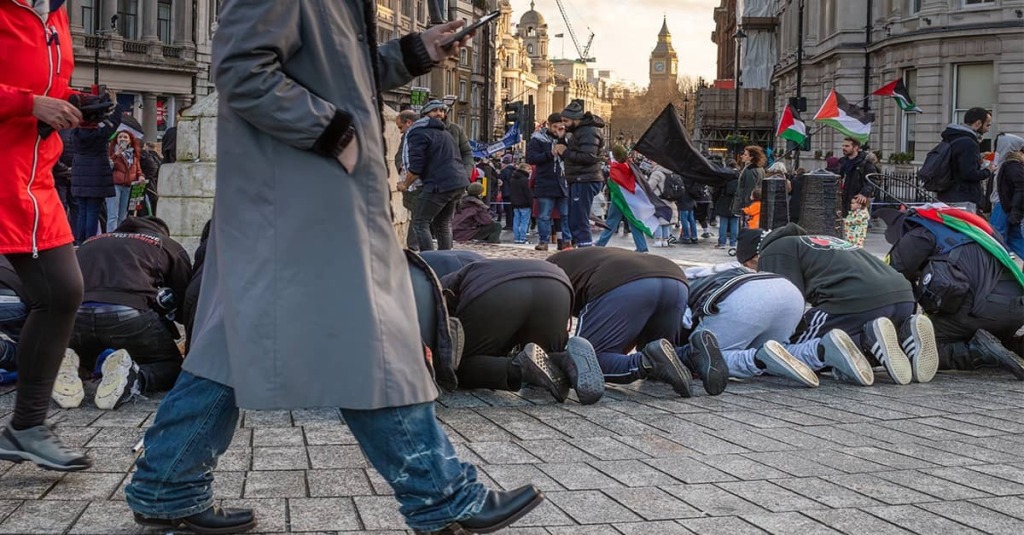 People kneel on a city street during a demonstration as others walk by, with multiple Palestinian flags visible and a large clock tower in the background.