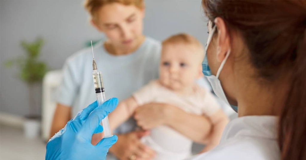 A healthcare worker holds up a syringe and approaches mother and baby in doctor's office.
