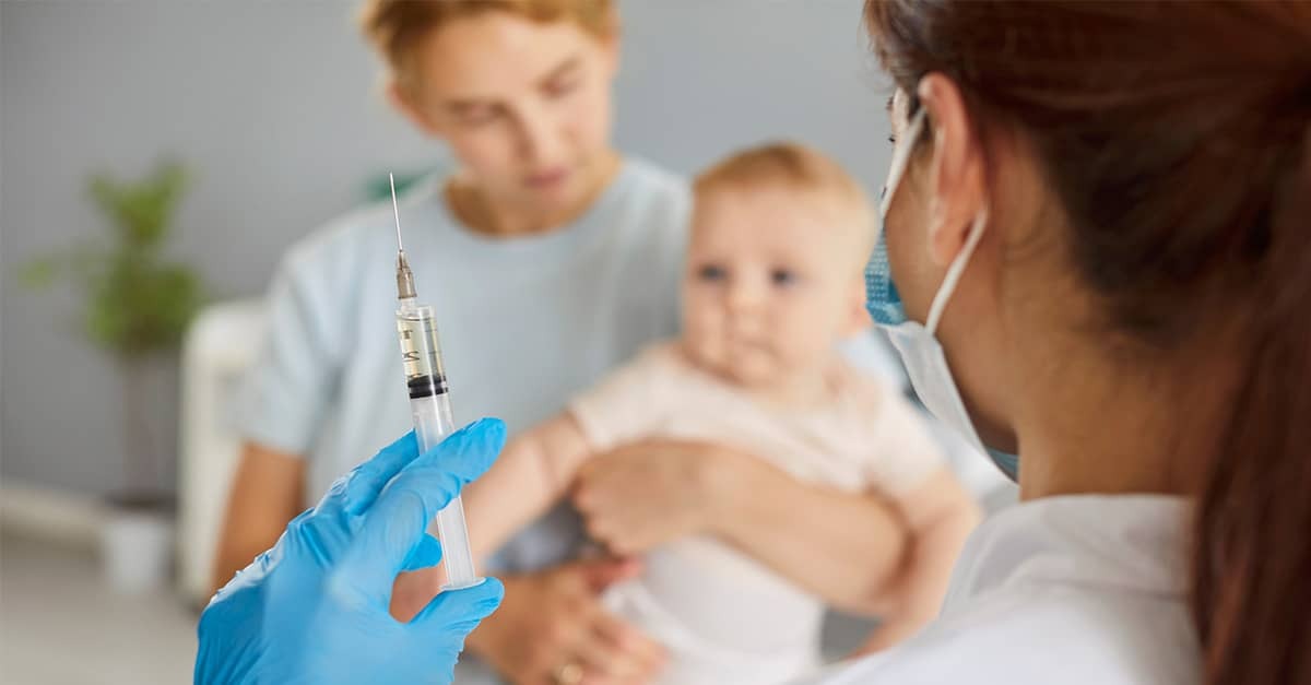 A healthcare worker holds up a syringe and approaches mother and baby in doctor's office.