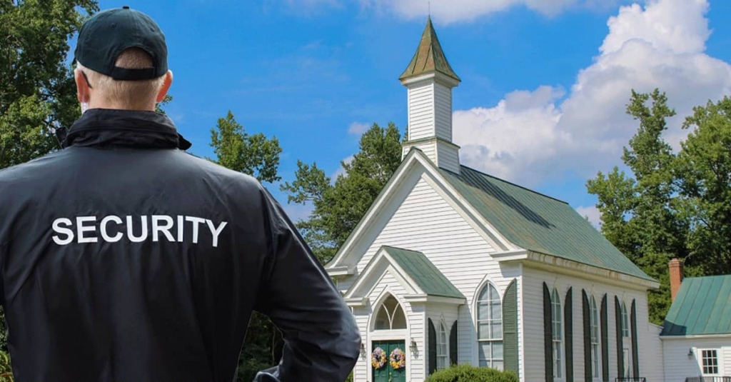 Security guard wearing a ‘SECURITY’ jacket stands outside a white church building, symbolizing protection of worship services.