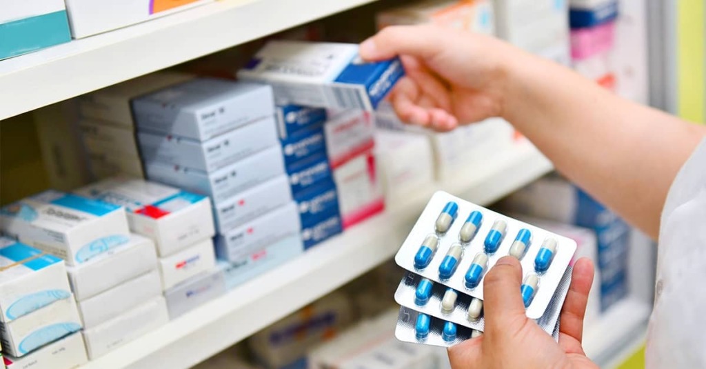 Hand holding blister packs of medication in a pharmacy aisle near prescription boxes.