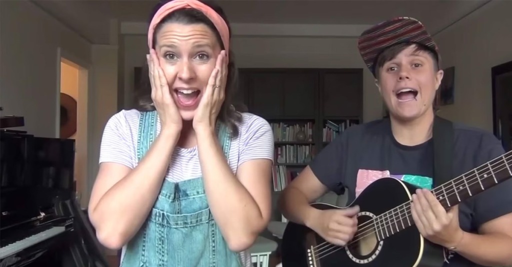 Ms. Rachel, a popular YouTuber and author who caters to toddlers, and Jules Hoffman, a non-binary musician, sing indoors during a children’s music video, with one playing a small guitar while the other reacts with an animated expression.