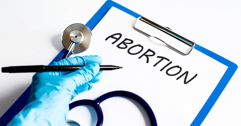 A gloved hand writes the word abortion on a medical clipboard beside a stethoscope.