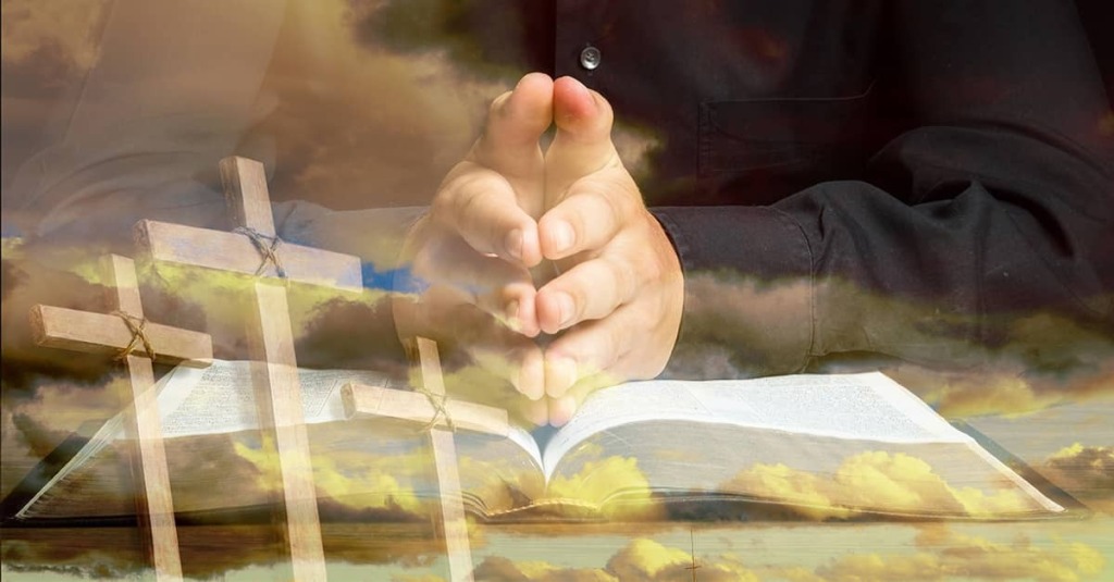 A man prays over a Bible, against the backdrop of three crosses, clouds and sunlight.