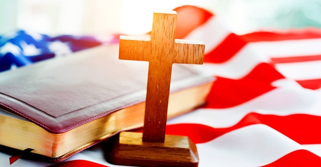 Wooden cross standing before a Bible laid over an American flag, symbolizing Christianity’s place in America’s public life and heritage.