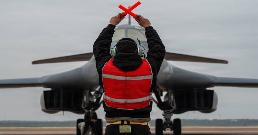 A military aircraft marshaller directs a B-1 bomber on a runway in support of Operation Epic Fury.