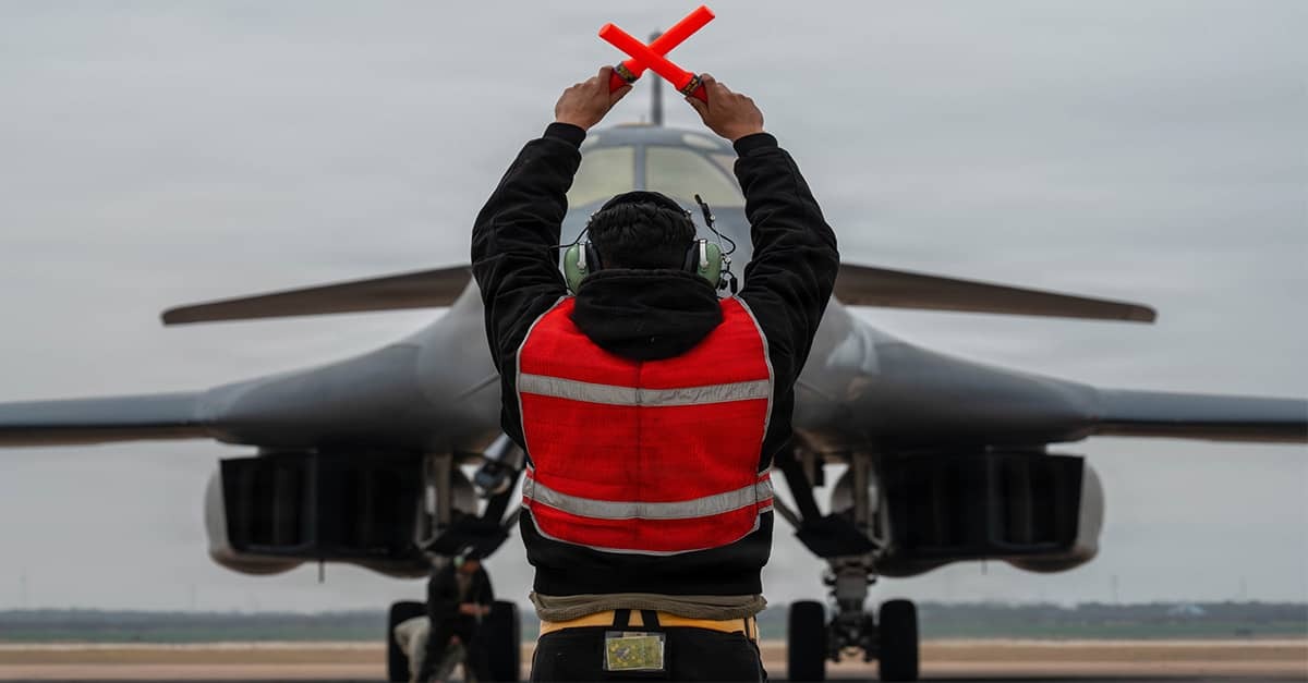 A military aircraft marshaller directs a B-1 bomber on a runway in support of Operation Epic Fury.