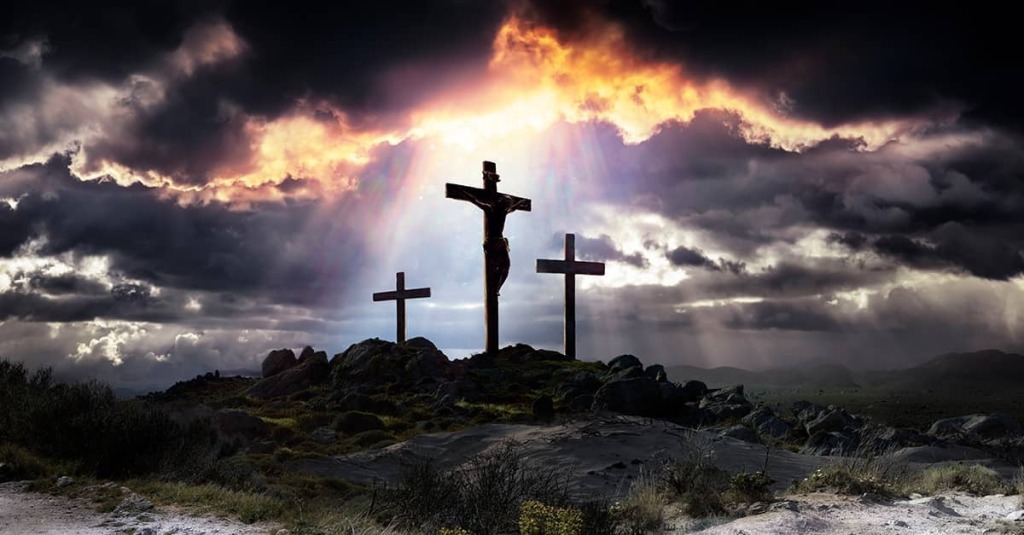 Silhouette of Jesus on the cross between two other crosses on a rocky hill as light breaks through dark storm clouds above Golgotha.