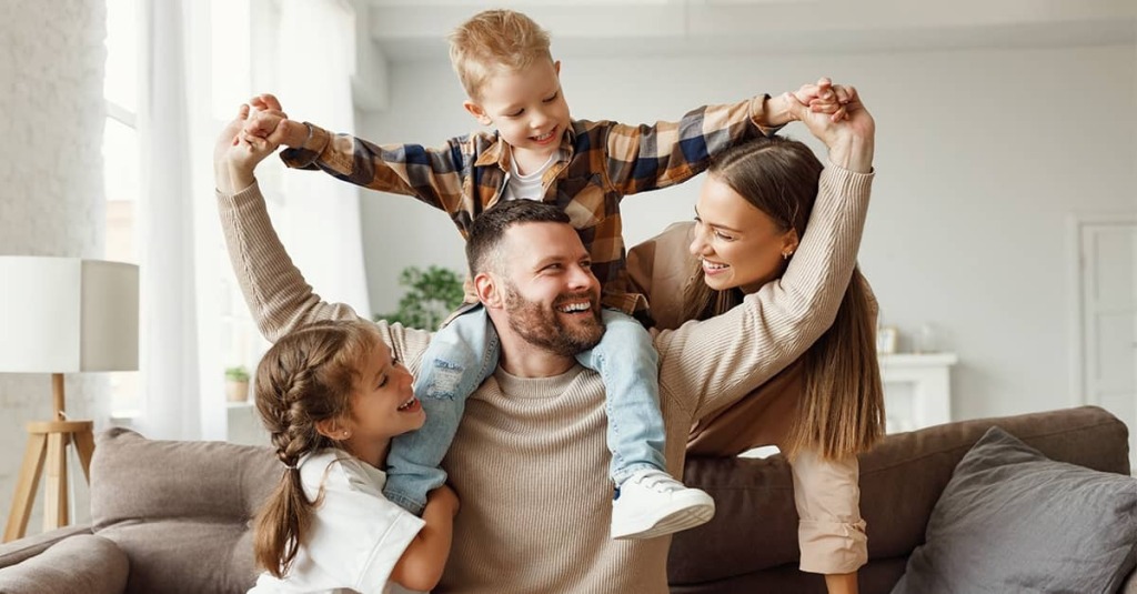 Smiling mother and father with a young son on the father’s shoulders and a young daughter beside them in a bright living room.
