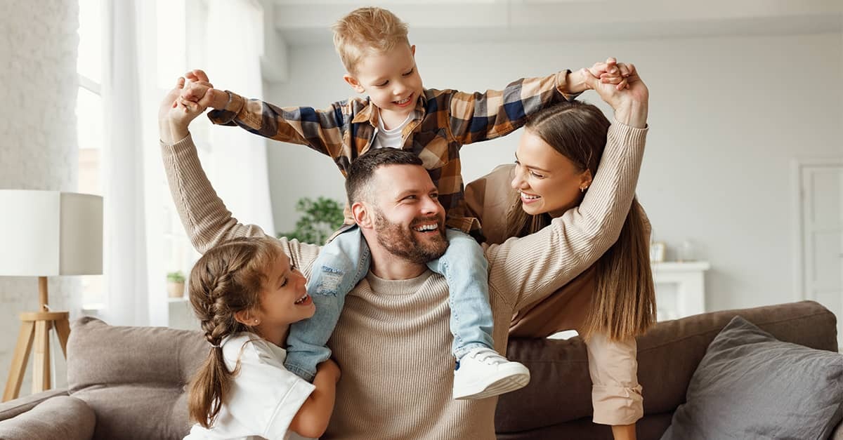 Smiling mother and father with a young son on the father’s shoulders and a young daughter beside them in a bright living room.