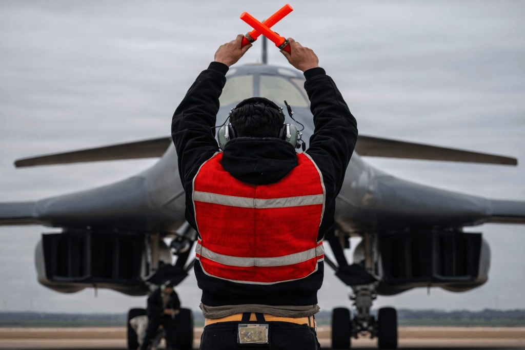 A military aircraft marshaller directs a B-1 bomber on a runway in support of Operation Epic Fury.