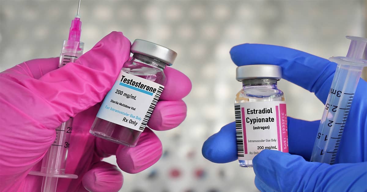 Gloved hands hold vials labeled testosterone and estradiol beside syringes, representing hormone drugs used in gender-transition treatments.