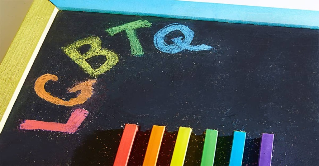 The letters "LGBTQ" are written on a school blackboard using rainbow colored chalk.