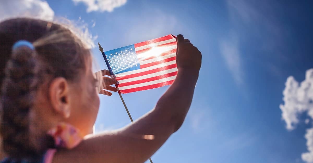 Girl holds up an American flag against sunlit sky.