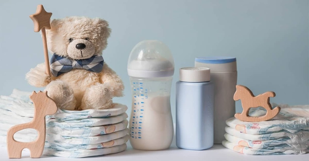 Teddy bear beside stacked diapers, baby bottles, and wooden nursery toys against a light blue background.