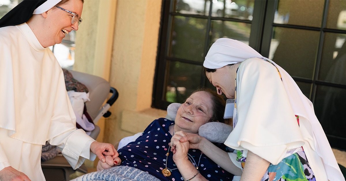 Two Dominican Sisters smile and hold hands with an elderly patient seated in a wheelchair in New York.
