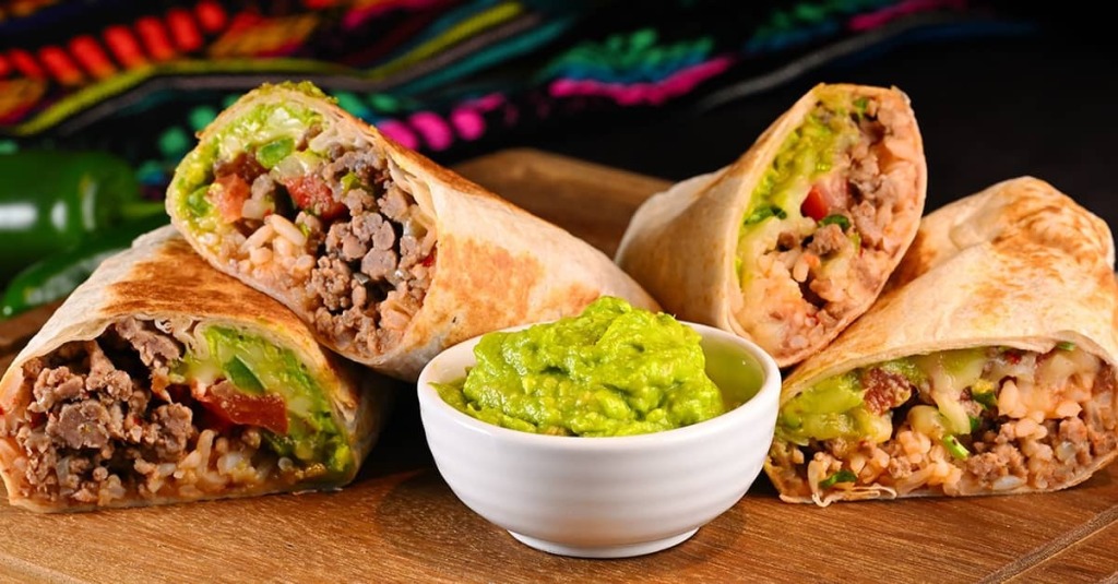 Four beef burritos filled with rice, avocado, tomatoes, and cheese arranged around a bowl of guacamole.