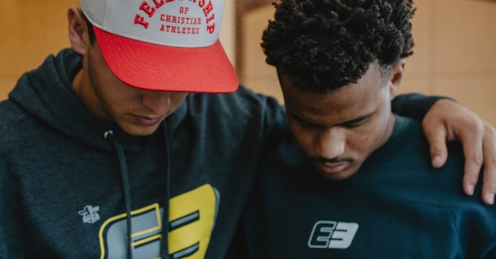 A young man wearing a hat that reads Fellowship of Christian Athletes prays with a student.