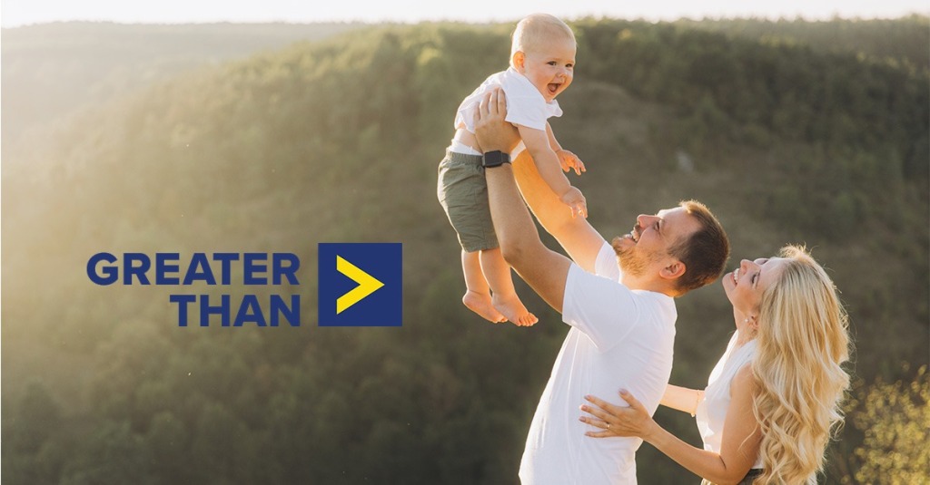 A mother watches as a father hoists their baby up in the air next to a logo for the "Greater Than" campaign.